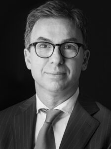 Zwart-wit portret van een man in pak en stropdas met bril, tegen een donkere achtergrond.