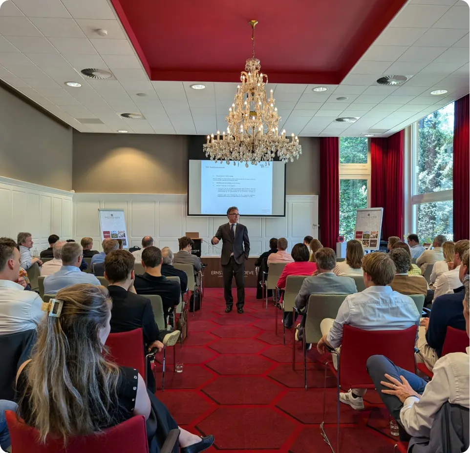 Frank geeft college aan een groep studenten in een conferentiezaal met een groot scherm en kroonluchter.